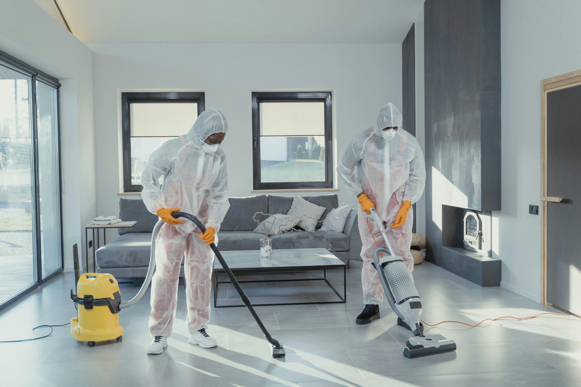Two workers in protective gear vacuum and disinfect a modern living room.